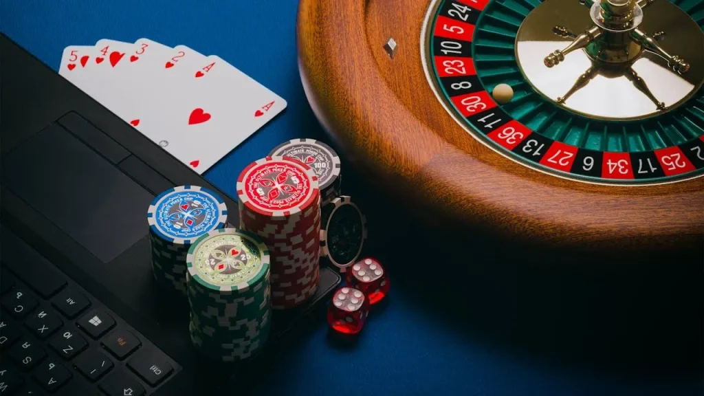 Gambling items on a blue surface, including a laptop keyboard, poker chips, red dice, playing cards (2 to 5 of hearts), and a roulette wheel.